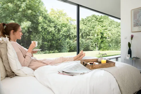 Frau liegt auf Bett, mit einer Tasse Tee vor einer Fensterfront